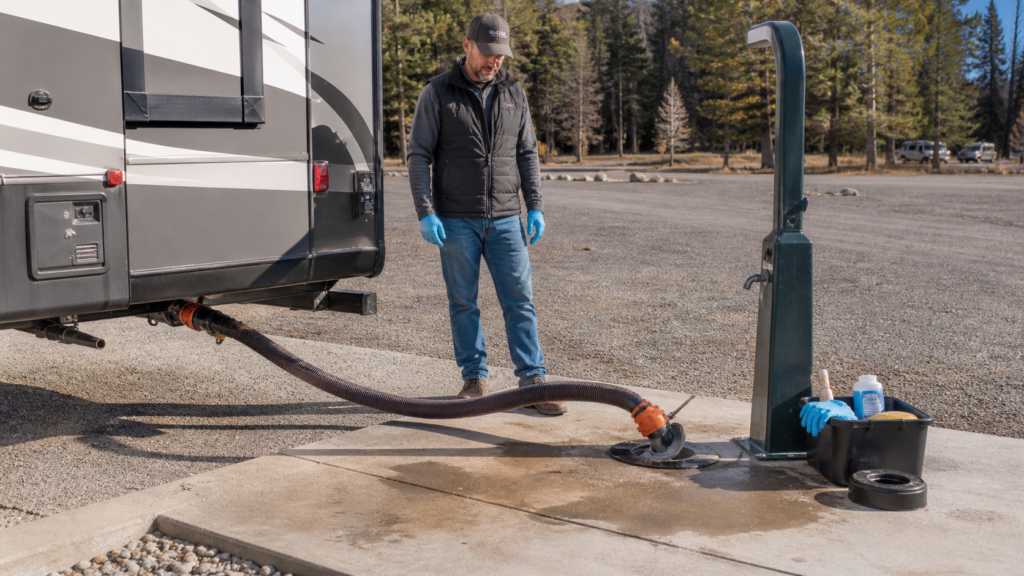 RV owner preparing to empty holding tanks at a campground dump station with a securely connected sewer hose and organized supplies.